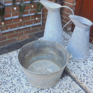 Op de afbeelding: Drie gegalvaniseerde metalen containers, twee kannen en een grote kom, allemaal met een rustieke, vintage uitstraling. De containers staan op een grijs en wit grind oppervlak.