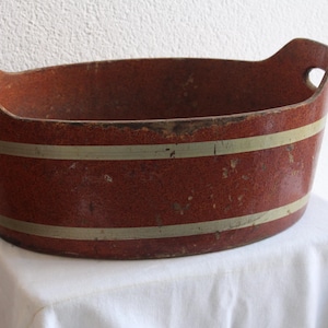 May include: An antique, oval-shaped, red and gold-striped wooden tub with handles. The tub has a distressed finish, suggesting age and use. The handles have heart-shaped cutouts. The tub is displayed on a white cloth.