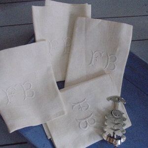 Puede incluir: Cuatro servilletas de lino blanco con iniciales bordadas "FB" en una fuente cursiva. Las servilletas están dobladas y dispuestas sobre una superficie azul con un sacacorchos plateado.