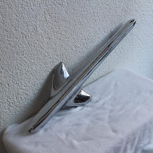 May include: A polished chrome car hood ornament, resembling a streamlined arrow or spear, rests on a white cloth. The ornament's design is sleek and reflective. The background features a textured, off-white wall. The object is likely vintage.