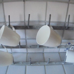 May include: A wall-mounted metal mug rack featuring a weathered gray finish with multiple vertical hooks. Four plain white ceramic mugs are hanging on the rack, positioned on different hooks across the two-tiered metal frame structure.