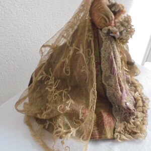 May include: A decorative doll wearing a layered gown featuring a gold-toned sheer lace veil with floral embroidery, a patterned fabric bodice in shades of gold and pink, and a ruffled skirt with intricate lace trim and small floral accents.