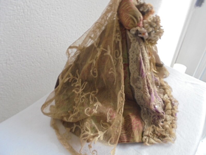 May include: A decorative doll wearing a layered gown featuring a gold-toned sheer lace veil with floral embroidery, a patterned fabric bodice in shades of gold and pink, and a ruffled skirt with intricate lace trim and small floral accents.