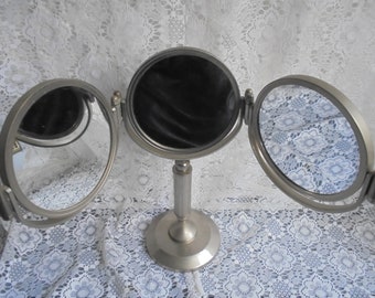 May include: A silver-toned, three-sided vanity mirror with a round base and a tall, slender column. The mirror is on a white lace tablecloth.