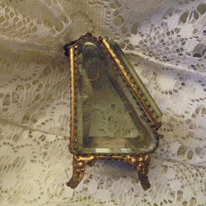 May include: A gold-toned triangular trinket box with a glass top and ornate detailing. The box is sitting on a white lace surface.