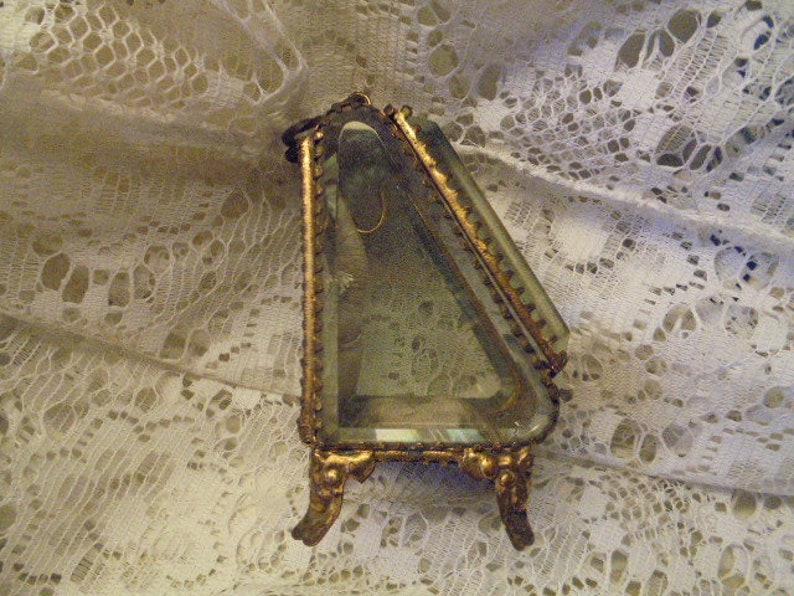May include: A gold-toned triangular trinket box with a glass top and ornate detailing. The box is sitting on a white lace surface.