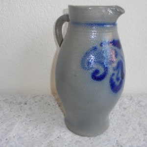 May include: A gray ceramic pitcher with a handle and spout. The pitcher features blue decorative swirls and a blue band around the top. The stoneware jug is likely used for serving beverages.