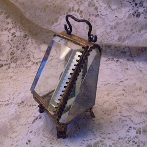 May include: A small, antique, glass display case with a gold-toned metal frame and a hinged lid. The case has a clear glass interior and sits on four small, decorative feet.