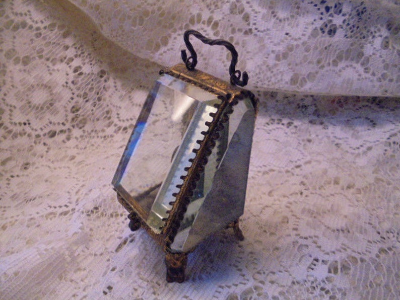 May include: A small, antique, glass display case with a gold-toned metal frame and a hinged lid. The case has a clear glass interior and sits on four small, decorative feet.