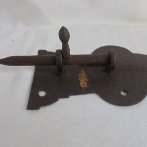 Op de afbeelding: Een roestige metalen deurgrendel met een puntige bout en een decoratieve plaat. De grendel is ontworpen om op een deur of poort te worden gemonteerd.