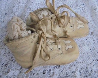 May include: A pair of antique baby shoes made of off-white leather with lace-up closures. The shoes have a worn and faded appearance, suggesting they are vintage.