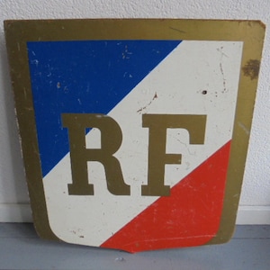 Puede incluir: Un letrero vintage de metal con forma de escudo y borde dorado. El escudo está pintado con los colores de la bandera francesa, azul, blanco y rojo. Las letras "RF" están en dorado en la sección blanca del escudo.
