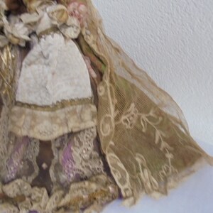 May include: A decorative doll wearing a layered gown made of tan and brown fabric with intricate lace overlays, purple ribbon accents, and a textured white apron. The doll features a flowing sheer veil with embroidered floral patterns.