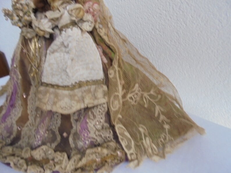 May include: A decorative doll wearing a layered gown made of tan and brown fabric with intricate lace overlays, purple ribbon accents, and a textured white apron. The doll features a flowing sheer veil with embroidered floral patterns.