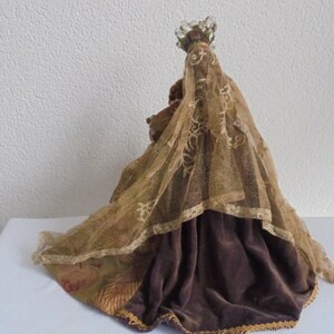 May include: A decorative figurine wearing a dark brown velvet skirt and a sheer, gold-toned lace veil draped over the head and shoulders. The figure is topped with a small, clear beaded crown ornament, and the fabric features intricate patterns.