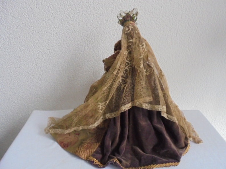 May include: A decorative figurine wearing a dark brown velvet skirt and a sheer, gold-toned lace veil draped over the head and shoulders. The figure is topped with a small, clear beaded crown ornament, and the fabric features intricate patterns.