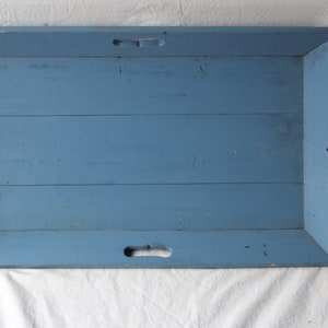 Pode incluir: Bandeja retangular de madeira azul com alças recortadas nas laterais. A bandeja tem uma aparência desgastada e rústica, com grão de madeira visível e um acabamento de pintura ligeiramente desbotado. Para servir ou exibir.