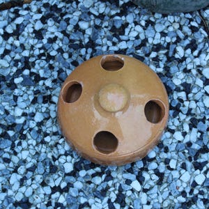 May include: A round, light brown ceramic object with four evenly spaced holes and a central knob. The object sits on a bed of small, blue and gray pebbles. The ceramic piece appears to be a decorative item.