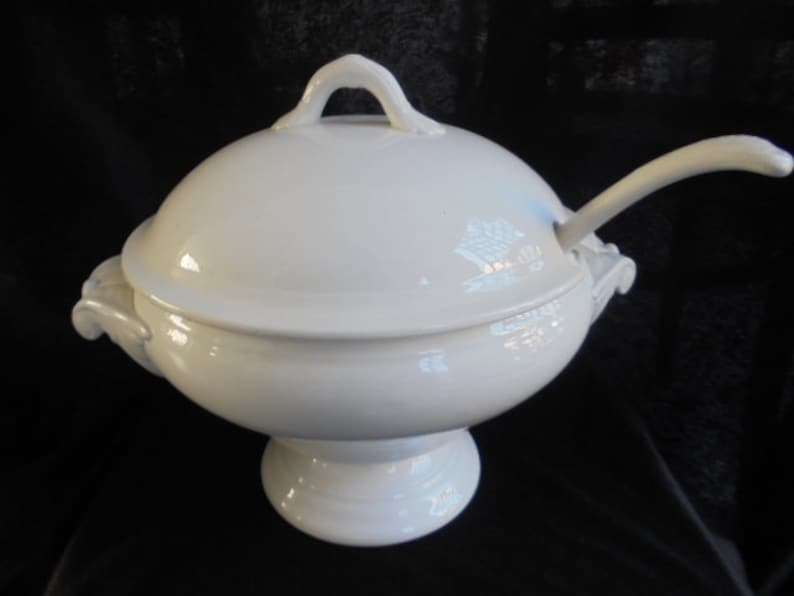 May include: White porcelain serving bowl with a lid and a ladle. The bowl has a round base and two handles on the sides. The lid has a knob on top.
