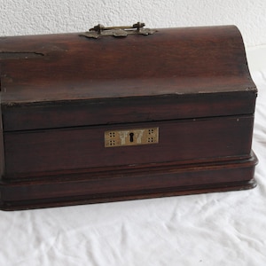 May include: A dark brown wooden box with a curved lid and a rectangular base. The box features a brass handle on top and a decorative brass keyhole plate on the front. The wood has a visible grain and a smooth, polished finish throughout the exterior.