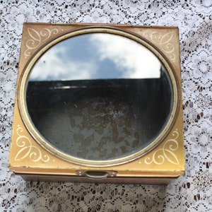 Op de afbeelding: Een vintage vierkante blikken doos met een rond, doorzichtig deksel. Het deksel heeft een gouden rand en een decoratief ontwerp rond de rand. De doos is geverfd in een lichtbruine kleur.