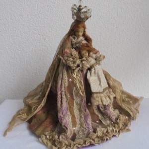 May include: A decorative doll featuring a figure with long reddish hair wearing a crown of clear beads, dressed in a layered gown of tan, gold, and purple fabric with lace trim. The figure holds a smaller doll in a cream-colored outfit.