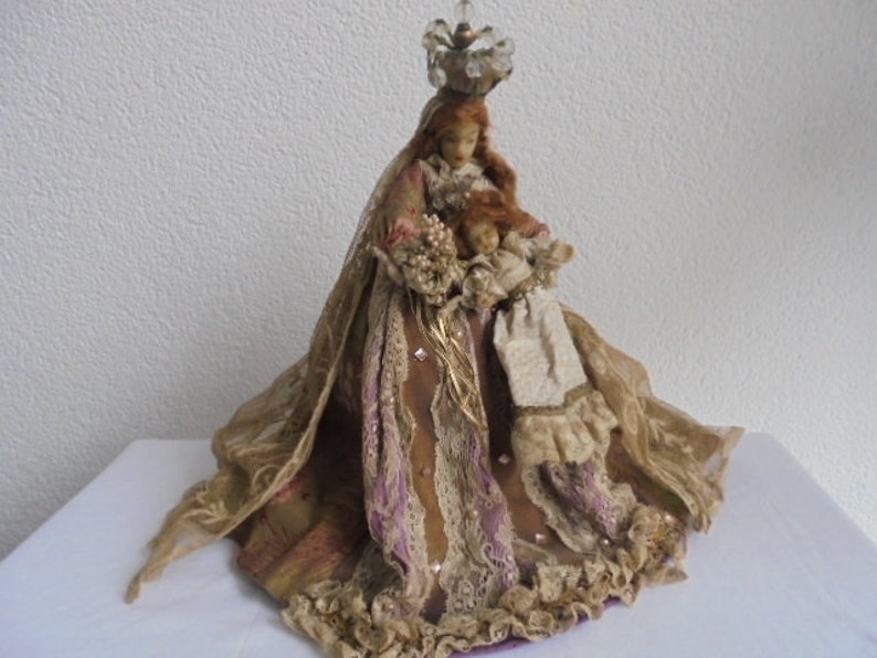 May include: A decorative doll featuring a figure with long reddish hair wearing a crown of clear beads, dressed in a layered gown of tan, gold, and purple fabric with lace trim. The figure holds a smaller doll in a cream-colored outfit.