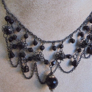 Op de afbeelding: Een zwarte kralenketting met een hangende zwarte kraal aan het einde. De ketting is gemaakt van meerdere kettingstrengen met zwarte kralen die aan elke streng zijn bevestigd.