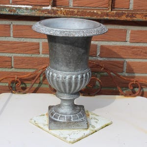 Op de afbeelding: Een verweerde, grijze metalen urn met een brede, decoratieve rand en een gegroefd lichaam. De urn staat op een vierkante basis bovenop een witte en gele tegel. De achtergrond is een rode bakstenen muur en een roestig metalen decoratief element.