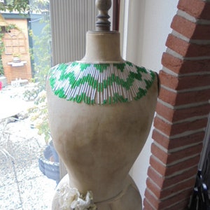 May include: A white dress form with a green and white beaded collar. The dress form is standing on a wooden floor.