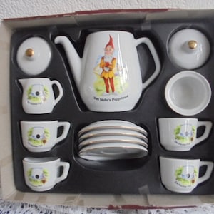 May include: A white porcelain tea set with gold trim, including a teapot, creamer, sugar bowl, six saucers, and three teacups. The teapot features a cartoon gnome illustration and the text "Van Nelle's Piggeme". The set is displayed in a cardboard box.