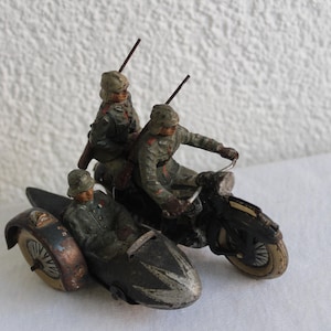 Könnte beinhalten: Ein Vintage-Spielzeugset mit drei Soldaten, die ein Motorrad mit Beiwagen fahren. Die Soldaten sind in Tarnfarben bemalt und halten Gewehre. Das Motorrad ist schwarz lackiert und hat einen Beiwagen in Braun-Schwarz.