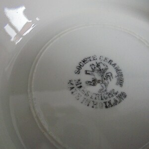May include: Close-up of the underside of a white ceramic plate with a black stamp that reads "Societe Ceramique  Manufacture  St. Amand  1920  Ikard" and a black image of a lion.