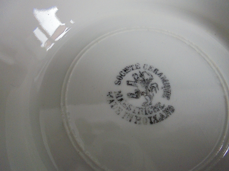 May include: Close-up of the underside of a white ceramic plate with a black stamp that reads "Societe Ceramique  Manufacture  St. Amand  1920  Ikard" and a black image of a lion.
