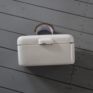 May include: A rectangular, off-white enamelled metal storage box with a hinged lid and a central latch. The box features visible dark wear along the edges of the lid and latch. A roll of brown adhesive tape is positioned behind the box.