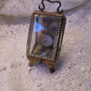 May include: A vintage pocket watch with Roman numeral markings on the face is displayed in a glass and metal case with a decorative metal frame.