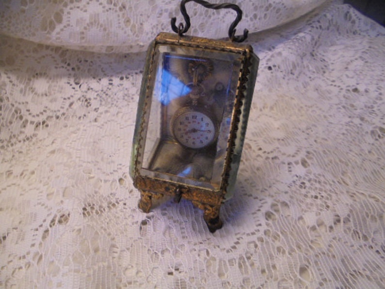 May include: A vintage pocket watch with Roman numeral markings on the face is displayed in a glass and metal case with a decorative metal frame.