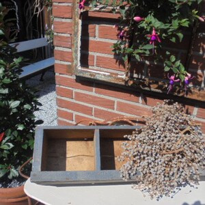 Op de afbeelding: Een blauwe houten doos met twee compartimenten, die op een witte tafel staat. De doos is leeg en heeft een rustieke uitstraling. De doos staat voor een bakstenen muur met een raamkozijn en een struik met roze bloemen.
