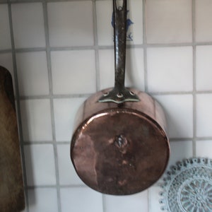 Könnte beinhalten: Ein Kupfertopf mit langem Griff, der an einer weißen gekachelten Wand hängt. Der Topf ist leer und hat eine glänzende Oberfläche.