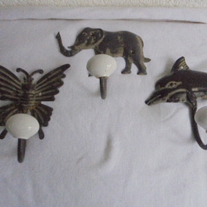 Op de afbeelding: Drie decoratieve wandhaken met porseleinen knoppen. De haken zijn gevormd als een vlinder, een olifant en een dolfijn.