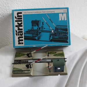 Puede incluir: Un juego de trenes modelo Märklin, con una caja azul que muestra el nombre de la marca y una representación de un paso a nivel. El juego incluye un paso a nivel en miniatura con un pequeño edificio verde, barreras y vías.