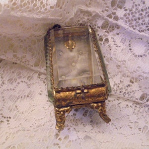 May include: A small, ornate, gold-colored, glass-topped trinket box with a clear glass lid and decorative feet. The box is sitting on a white lace surface.