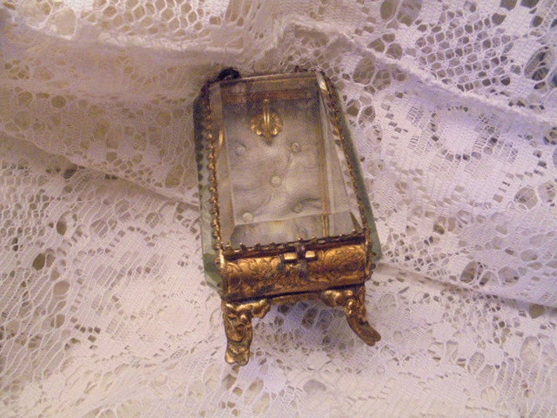 May include: A small, ornate, gold-colored, glass-topped trinket box with a clear glass lid and decorative feet. The box is sitting on a white lace surface.