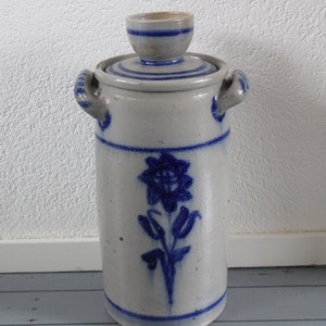 Op de afbeelding: Een hoge, witte keramische pot met een blauw en wit bloemenmotief. De pot heeft een deksel met een kleine, ronde knop bovenop. De pot is versierd met een blauw en wit bloemenmotief.