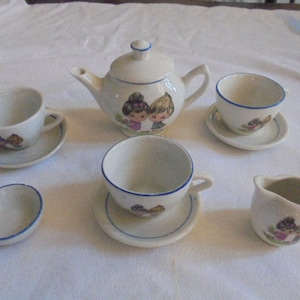 May include: A white porcelain children's tea set with a blue trim. The teapot features a cartoon illustration of a girl and boy. The set includes a teapot, creamer, sugar bowl, and two teacups and saucers.
