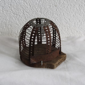 May include: A dome-shaped wire cage with a rusted brown finish, featuring a circular metal top plate and vertical metal slats. The cage is mounted onto a thick, weathered wooden base with a rough, uneven texture and a visible metal fastener.