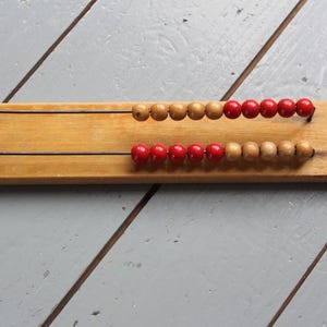 May include: A vintage wooden abacus with two rows of beads. The beads alternate between natural wood and red, strung on thin wires. The abacus is set against a gray wooden surface.