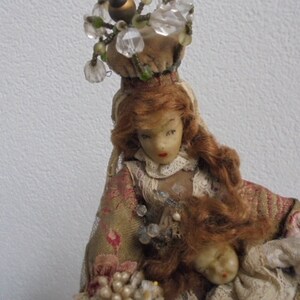 May include: A doll with auburn hair wearing a tan floral patterned garment with lace trim and a crown-like headpiece adorned with clear beads. The doll holds a smaller doll figure with a pale face and red lips, featuring intricate beadwork.