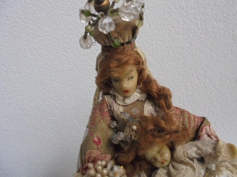 May include: A doll with auburn hair wearing a tan floral patterned garment with lace trim and a crown-like headpiece adorned with clear beads. The doll holds a smaller doll figure with a pale face and red lips, featuring intricate beadwork.