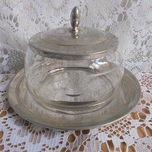 Könnte beinhalten: Eine versilberte Glasbutterdose mit einem gewölbten Deckel. Die Dose hat einen klaren Glasboden und einen versilberten Rand. Der Deckel hat einen versilberten Knopf.